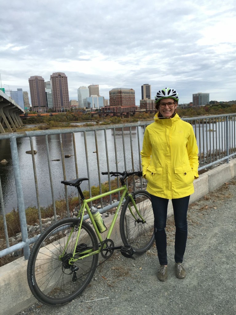 Nicole Parker in a yellow rain jacket and bicycle helmet beside her Surly bike on a James River path, with the Richmond skyline behind her
